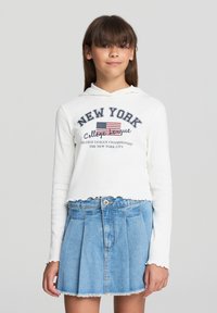 Felpa bianca con maniche lunghe e stampa "NEW YORK" e bandiera, abbinata a una gonna di jeans blu con pieghe e orlo sfrangiato.