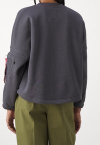 Tmavě šedá mikina s kulatým výstřihem, volným střihem, elastickými manžetami a boční kapsou s červenou štítkem. Nošena přes olivově zelené kalhoty.