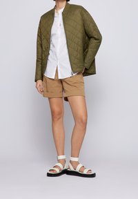 Olivově zelená prošívaná bomber bunda se zipem, bílá košile s knoflíky, hnědé šortky a bílé sandály s pásky a černou podrážkou.