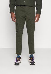 Man som bär olivgröna cargobyxor och flerfärgade sneakers står framför en enkel ljusgrå bakgrund.