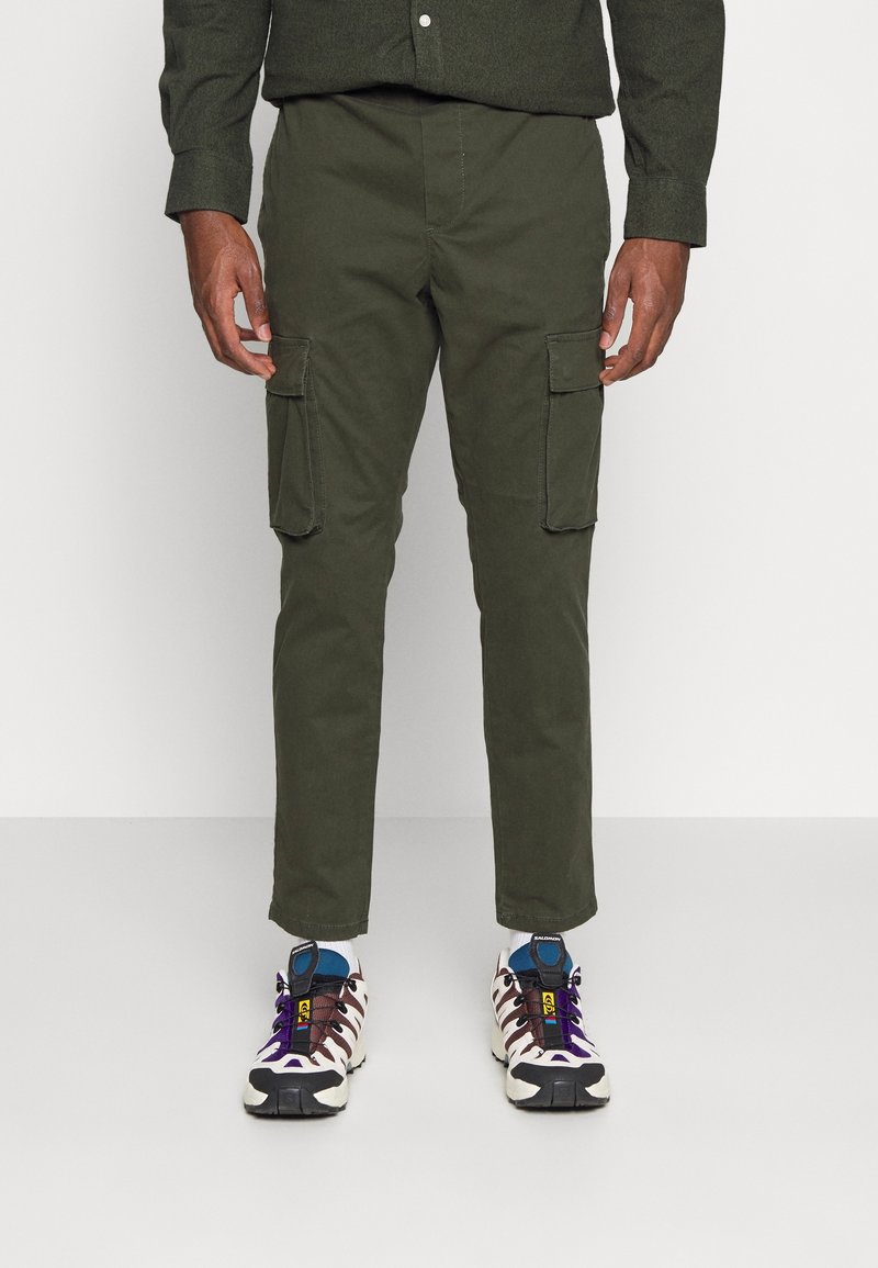 Man som bär olivgröna cargobyxor och flerfärgade sneakers står framför en enkel ljusgrå bakgrund.