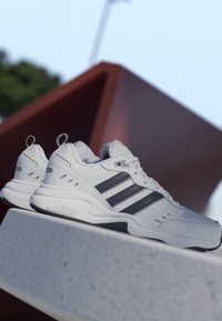 Baskets blanches Adidas avec des bandes noires posées sur une surface en pierre, sur fond de sculpture moderne floue et ciel clair.