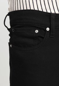 Svarta denimjeans med en åtsittande design, fem fickor och silverfärgad detaljer. Tyget har en slät, något texturerad yta.