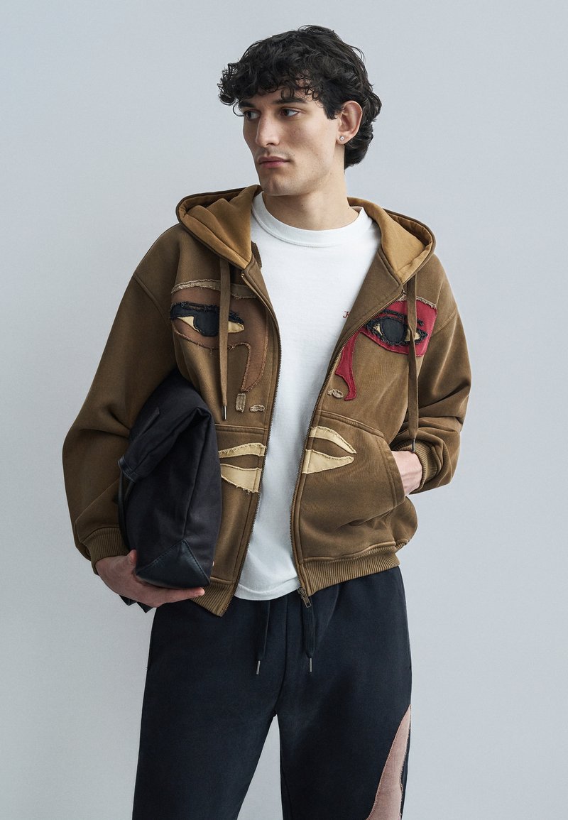 Καφέ φούτερ με φερμουάρ και abstract κεντήματα προσώπου σε κόκκινο και μαύρο, συνδυασμένο με λευκό μπλουζάκι και σκούρο παντελόνι. Κρατάει μια μαύρη τσάντα.