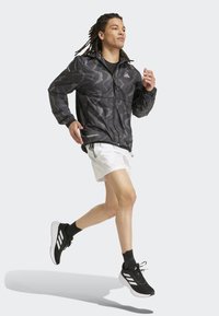 Schwarze Camouflage-Windjacke mit Reißverschluss vorne, elastischen Bündchen und Kordelzug-Saum, kombiniert mit weißen Shorts und schwarzen Sportschuhen.