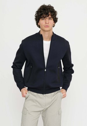 Giacca zip-up blu realizzata in tessuto liscio, con due tasche laterali con cerniere nere e un colletto arrotondato. Indossata sopra una camicia bianca.