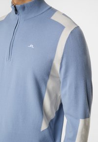 Ljusblå tröja med hög krage, utrustad med vita vinkelaksenter och en halvmåne dragkedja fram. Mjuk textur, logotyp på bröstet.