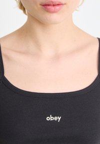 Svart ribbet tanktop med rund halslinning og liten brodert "obey"-logo i gull på forsiden.