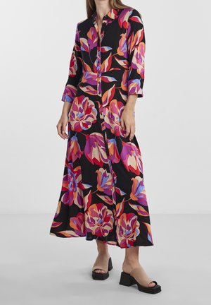 Kobieta ubrana w czarną sukienkę maxi z długim rękawem, ozdobioną żywymi, fioletowymi, czerwonymi i pomarańczowymi kwiatowymi wzorami oraz beżowe sandały na słupku z odkrytymi palcami.