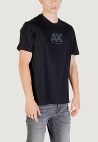 T-shirt en coton noir avec un col rond et des manches courtes, arborant un logo "AX" gris foncé sur la poitrine et une texture lisse.
