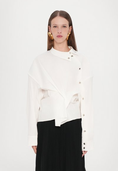 Chemise blanche à manches longues avec drapés asymétriques, col et accents de boutons argentés. Associée à une jupe plissée noire.