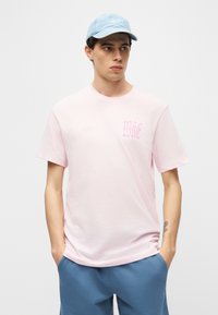 Νεαρός άνδρας με ανοιχτό μπλε καπέλο, ανοιχτό ροζ μπλουζάκι με σχέδιο και μπλε σορτς, στέκεται με τα χέρια στις τσέπες μπροστά σε λευκό φόντο.