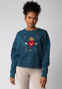 Femme portant un sweat-shirt teal avec un cœur flamboyant rouge et un motif floral, associé à un pantalon côtelé beige clair, se tenant devant un fond uni.