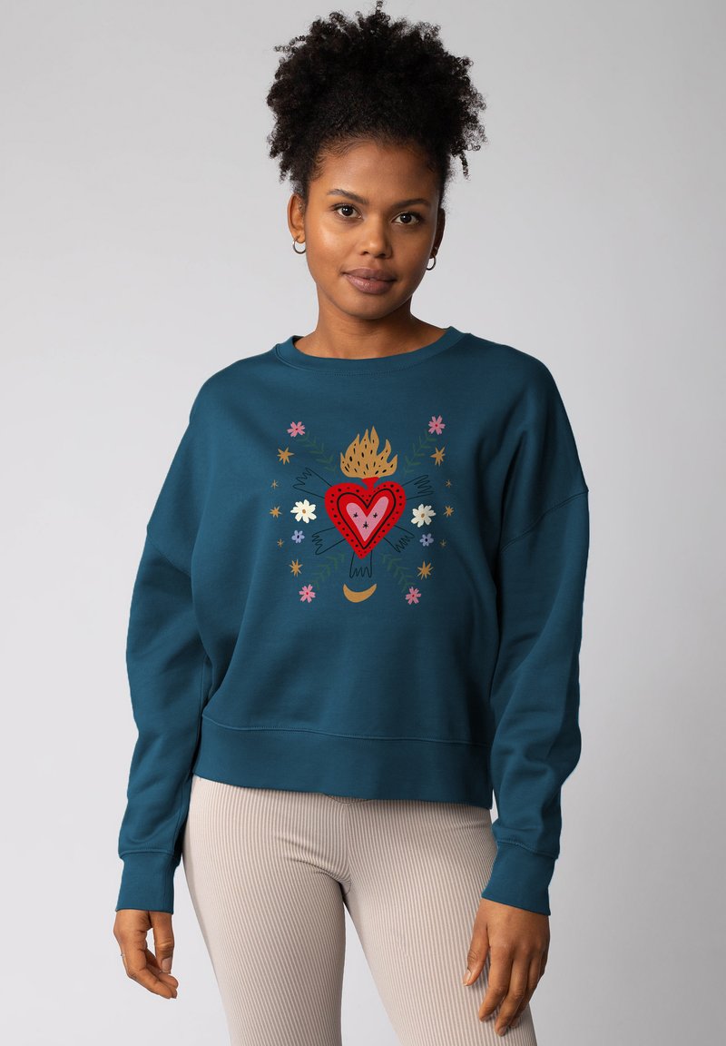 Femme portant un sweat-shirt teal avec un cœur flamboyant rouge et un motif floral, associé à un pantalon côtelé beige clair, se tenant devant un fond uni.