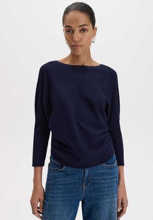 Frau mit dunklem, zurückgebundenem Haar, die ein dunkelblaues Langarmshirt und blaue Jeans trägt, steht vor einem weißen einfarbigen Hintergrund.