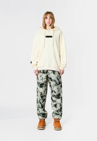 Sudadera crema con un parche de logo en el frente, combinada con pantalones de rayas grises y verdes; usados con botas marrones. Corte relajado y textura suave.