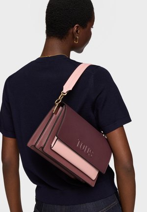 Persona con camisa azul marino oscuro lleva un bolso de hombro de cuero burdeos y rosa con "TOUS" grabado en la solapa.