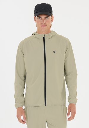 Young man wearing beige zip-up hooded jacket and matching pants with black cap, standing against plain white background.