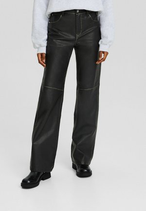 Person wearing black faux leather wide-leg pants, black boots, and a light grey long-sleeve top, standing against a neutral background.