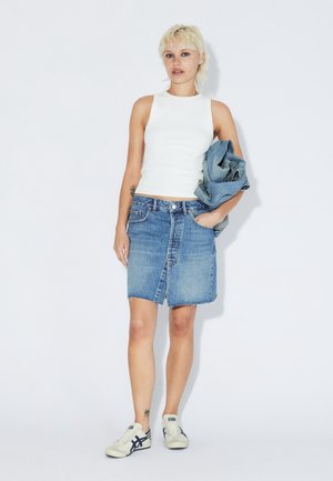 Ein hellblauer Jeansrock mit ausgefranstem Saum, kombiniert mit einem weißen ärmellosen Top und cremeweißen Turnschuhen, dazu eine Jeansjacke in der Hand.