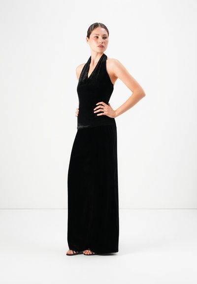 Woman standing with hands on hips, wearing a sleeveless black velvet halter top and matching wide-leg pants with black sandals.