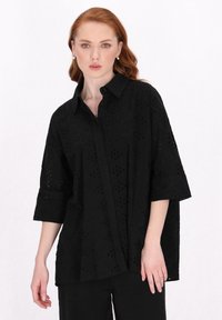 Schwarze Bluse mit kurzen Ärmeln und strukturiertem Kragen, ausgestattet mit einem texturierten Überlagerungsmuster, Knopfverschluss vorne und einem lockeren Schnitt.