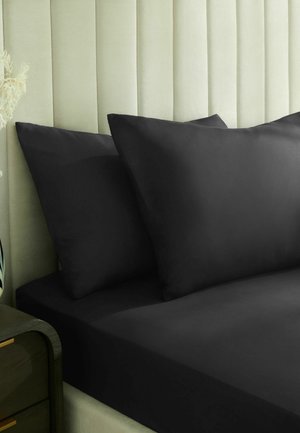 Schwarze Kissenbezüge und Spannbettlaken auf einem Bett mit einem hellgrünen gepolsterten Kopfteil und einem dunkelgrünen Nachttisch.