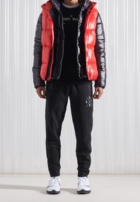 Gilet imbottito rosso e nero sopra una t-shirt nera, abbinato a pantaloni della tuta neri con logo e scarpe da ginnastica bianche.