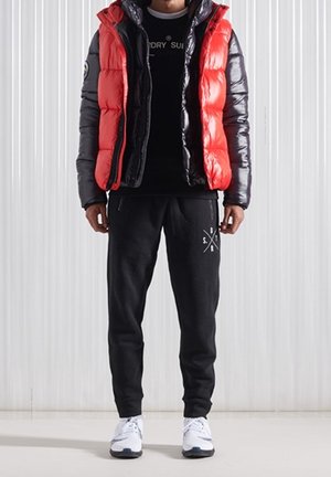 Gilet imbottito rosso e nero sopra una t-shirt nera, abbinato a pantaloni della tuta neri con logo e scarpe da ginnastica bianche.
