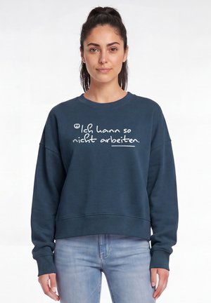 Junge Frau mit dunklen Haaren im Pferdeschwanz, die einen marineblauen Pullover mit dem deutschen Text "Ich kann so nicht arbeiten" und hellblaue Jeans trägt.