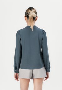 Kvinne med blond hestehale iført en langermet mørkegrå bluse og lysegrå shorts, som står med ryggen mot en ensfarget hvit bakgrunn.