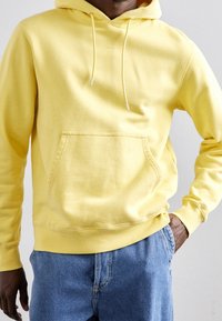 Felpa gialla in cotone con tasca a marsupio, cappuccio con cordino e polsini a coste, abbinata a jeans in denim azzurro chiaro.