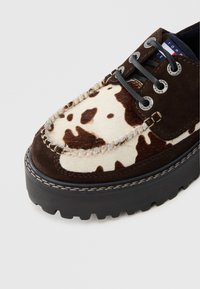 Brun og hvidt cowhide-mønstret sko med en kraftig sort platform sål, der har mørke snørebånd og detaljeret syning omkring kanterne.