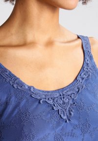 Modrý top bez rukávů s krajkovým výstřihem a květinovým vzorem. Materiál je lehký a strukturovaný, s kulatým límcem a tenkými ramínky.