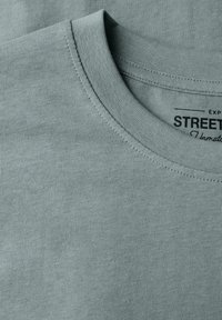 Světle modré tričko s kulatým výstřihem, viditelnými švy a částečně viditelným černým potiskem uvnitř límce.