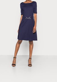 Mörkblå kortärmad klänning med figurnära liv, utsvängd kjol och metallisk bälteaccent i midjan. Slät tygtextur.