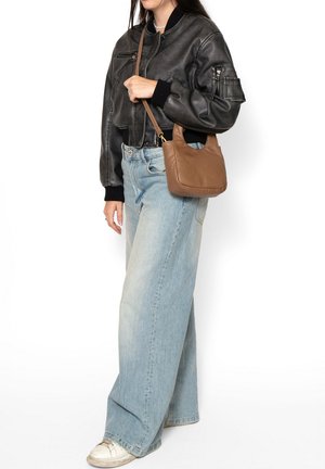Donna con giacca bomber in pelle nera e jeans larghi azzurri, con una piccola borsa a tracolla marrone in pelle, in piedi di lato.