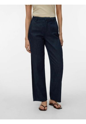 Femme portant un jean droit bleu marine, un haut beige à manches courtes, et des sandales plates marron, debout devant un fond blanc uni.