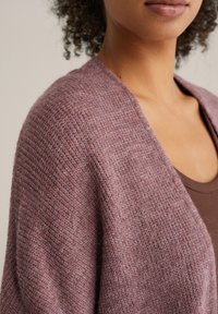 Cardigan supradimensionat dintr-un tricot moale și texturat, de culoare mov pal, cu un design minimalist și cu partea din față larg deschisă. Bluza maro stratificată este vizibilă dedesubt.