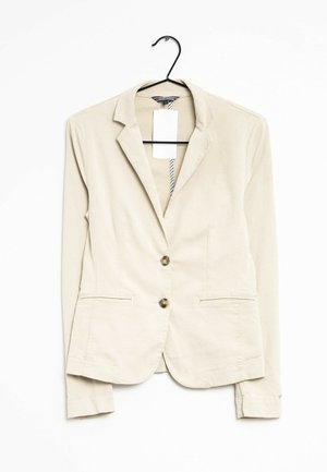 Beiger Blazer mit zwei Knöpfen und zwei Vorderaschen, der an einem schwarzen Bügel vor weißem Hintergrund hängt.