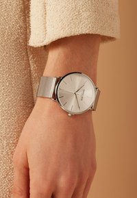 Silberner Armbanduhr mit einem Mesh-Armband, runder polierter Gehäuse und einem hellsilbernen Ziffernblatt. Dünne schwarze Indizes und Zeiger zeigen die Zeit an.