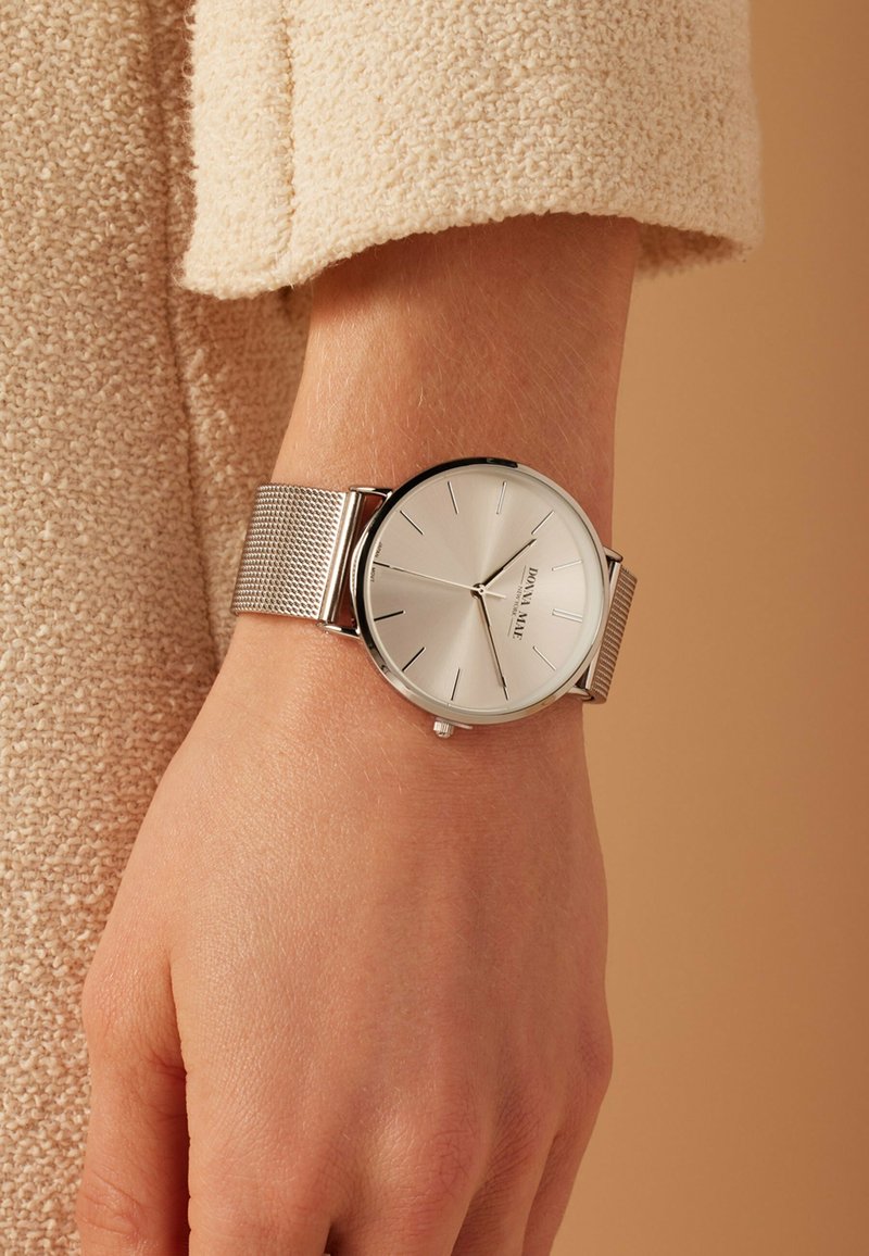 Silberner Armbanduhr mit einem Mesh-Armband, runder polierter Gehäuse und einem hellsilbernen Ziffernblatt. Dünne schwarze Indizes und Zeiger zeigen die Zeit an.