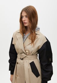 Beżowy trencz z czarnymi materiałowymi rękawami. Posiada podwójne guziki, pasek oraz kieszeń naszywaną na lewym rękawie.