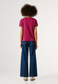 Magenta trumpa rankovė t-shirt, derinama su plačiomis mėlynomis džinsinėmis kelnėmis. Marškiniai turi laisvą siluetą; kelnės pasižymi klasikinio penkių kišenių dizainu.