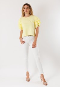 Top corto in cotone giallo con maniche a strati e volant, abbinato a jeans bianchi aderenti e sandali beige con tacco a strappo. Design semplice.