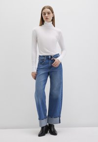 Weißes Langarm-Turtleneck, blaue weite Jeans mit umgeschlagenen Säumen, kombiniert mit schwarzen Stiefeletten, vor einem hellgrauen Hintergrund.