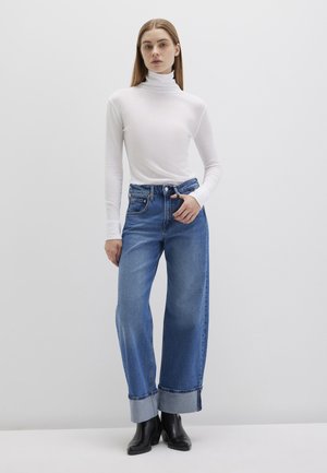 Weißes Langarm-Turtleneck, blaue weite Jeans mit umgeschlagenen Säumen, kombiniert mit schwarzen Stiefeletten, vor einem hellgrauen Hintergrund.