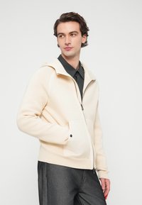 Φωτεινή μπεζ φούτερ με φερμουάρ, κουκούλα με κορδόνι και μπροστινές τσέπες. Διαθέτει ριμπ μανσέτες και τελείωμα, συνδυασμένο με σκουρόχρωμο πουκάμισο με γιακά.