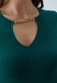 Teal ribbet topp som har V-hals og detaljer med gullkjede i halsen, som fremhever teksturert stoff og strukturert design.