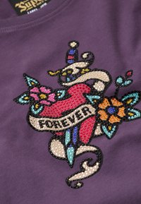 Tela de algodón morada con un diseño colorido que incluye un corazón rojo, flores y un banner ornamentado que dice "POR SIEMPRE", decorado con lentejuelas.